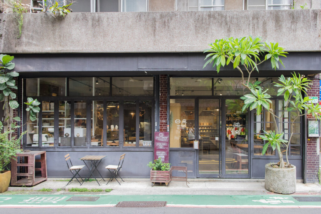 筑子 CHUZI Dialogue 永康街店面，深灰色外觀搭配大片玻璃窗與綠色植栽，門口設有戶外桌椅，呈現低調自然的老屋咖啡店氛圍。